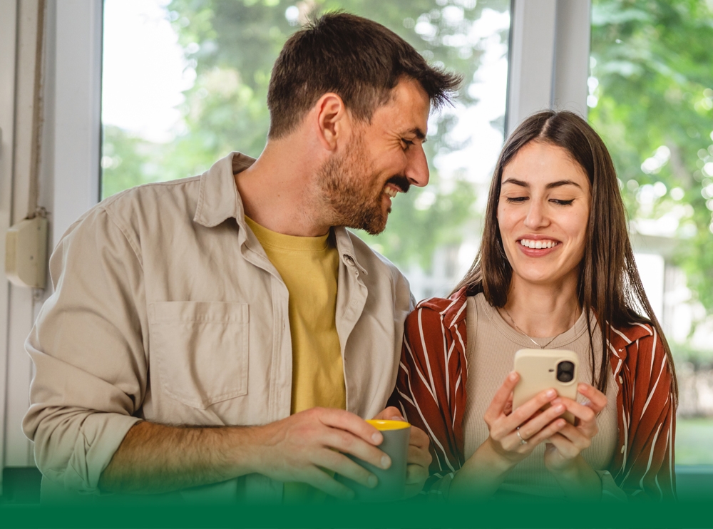 A couple at the kitchen sink, looking at their cell phone and laughing.